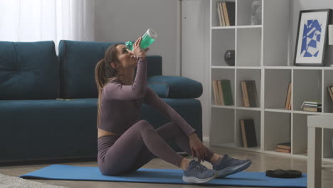 young-sportswoman-is-drinking-water-at-break-during-home-fitness-training-sitting-on-floor-in-living-room-workout-for-body-shaping